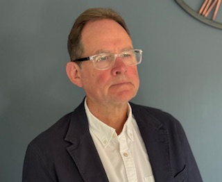 Person in a dark blazer over a white button-up shirt standing against a plain background, with part of a clock visible in the top right corner.