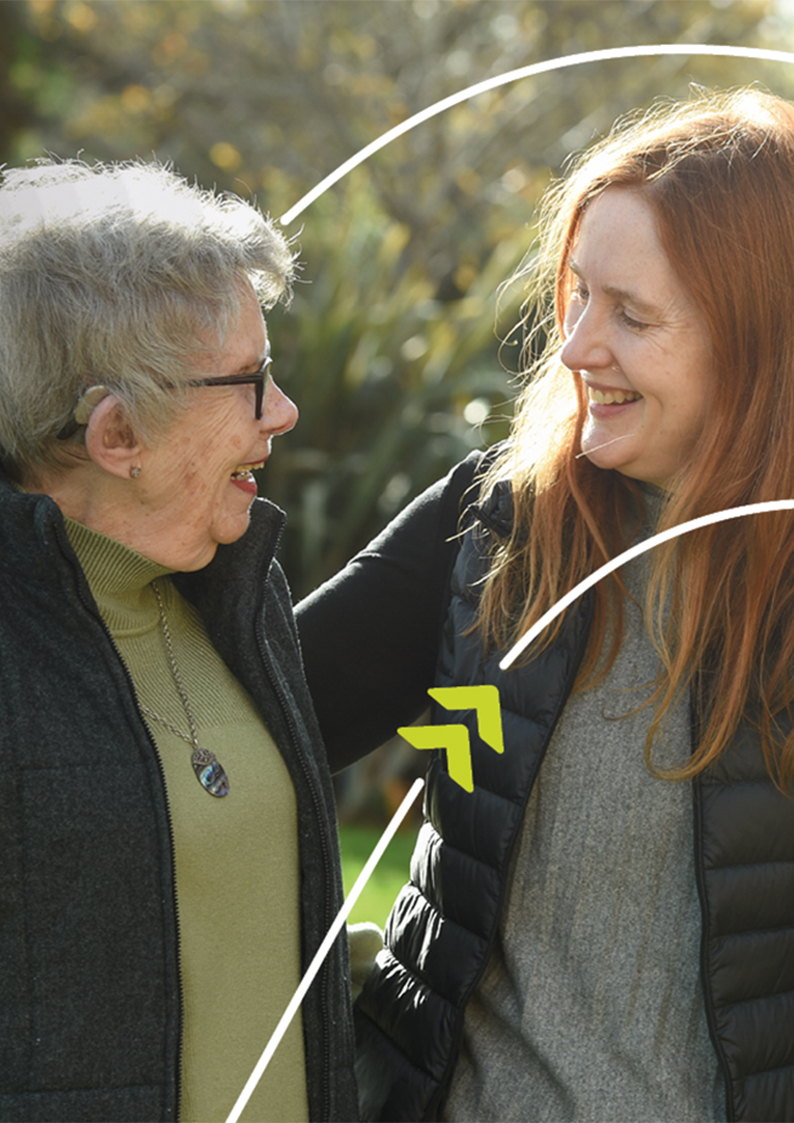 Two people standing together outdoors in a garden, with one person placing an arm around the other's shoulder.