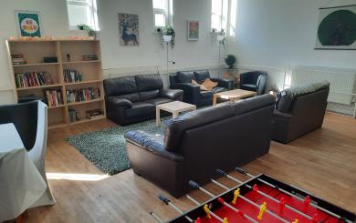 An daytime photo of the Community Living Room, showing a table and chairs, book case, sofas, coffee tables, and football table.