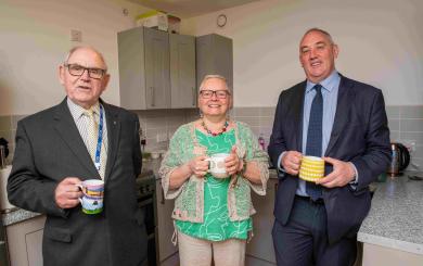 Three people standing up in a kitchen, smiling and holding mugs