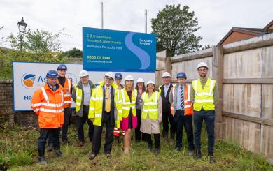 A group of people wearing hard hats and hi-vis from Sanctuary and the Renfrewshire Council stood on-site in front of signage for the 2 & 3 bedroom apartments which will be available for social rent