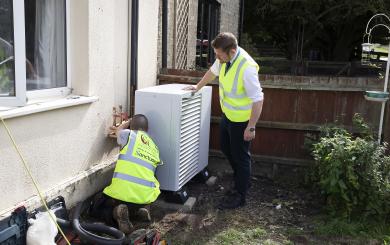 Two white, male Sanctuary staff members wearing high vis jackets are outside a building fitting a swaffham heat pump