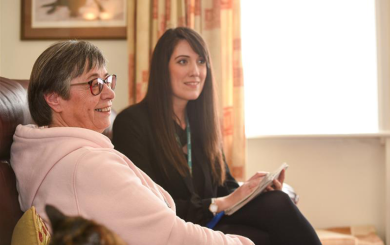 Alt text: A Sanctuary customer, a middle-aged woman with short grey hair and glasses, wearing a light pink hoodie, sits on a brown leather sofa, smiling. Next to her, a Sanctuary Housing Officer, a younger woman with long dark hair, dressed in a black blazer and wearing a lanyard, holds a notepad and pen. They are in a well-lit living room with cream curtains, a framed picture on the wall, and a cat partially visible in the foreground.