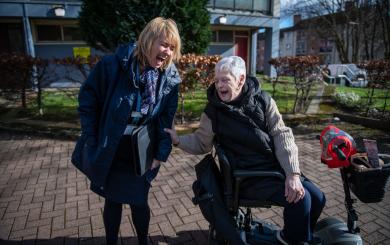 A smiling woman in a blue coat stands next to an elderly woman in a mobility scooter. They are both laughing and engaged in a joyful conversation outdoors, with a backdrop of a building and neatly trimmed bushes. 