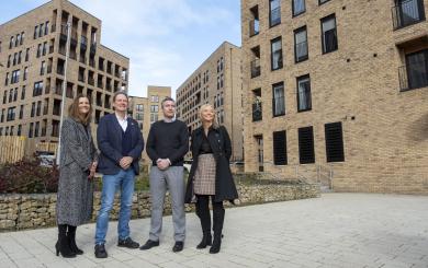 Four people standing in an outdoor courtyard area surrounded by modern multi-storey brick apartment buildings, with landscaped stone planters and paved walkways