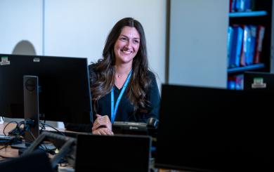 A woman with long, wavy hair is sitting at a desk in an office environment, smiling at the camera. She is positioned in front of multiple computer monitors, with a blue lanyard around her neck. 
