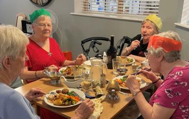 A group of four elderly women sitting around a table enjoying a meal together. They are wearing colorful paper crowns and are engaged in conversation.