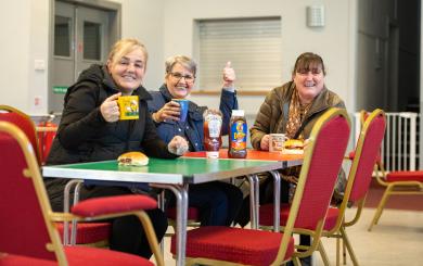 Three women are sitting at a table in a casual setting, smiling towards the camera. Each woman holds a mug, and there are bottles of sauce and sandwiches on the table.