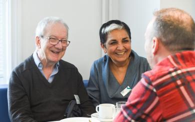 Three people sit around a small table, engaged in conversation with warm smiles. Cups and plates are on the table in a bright indoor setting.