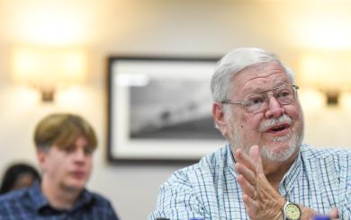 An older individual wearing glasses and a checkered shirt is seated indoors, with one hand slightly raised in a gesture as if speaking to someone out of frame. The person also wears a wristwatch on the left wrist. In the background, another individual is visible but blurred, appearing to listen to the conversation.