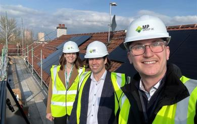 Donna Williams, Group Director of Strategy and Sustainability, is pictured with colleagues during a rooftop site visit. The group is standing on a scaffolded roof fitted with rows of solar panels, wearing hard hats and high visibility vests. Residential rooftops and a partly cloudy sky are visible in the background.