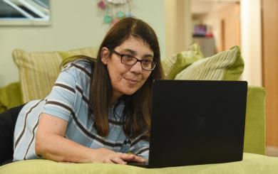 A woman with long brown hair and glasses is lying on a green couch, intently looking at a laptop in front of her. The background features a softly lit room with a hint of decor, suggesting a cozy home environment.