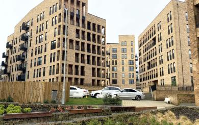 Modern mid-rise apartment buildings built with light brick surround a landscaped courtyard with terraced stone planters, benches, and small shrubs. Several parked cars sit on a grassy area and paved drive between the buildings under an overcast sky
