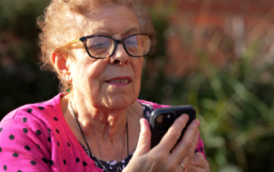 An older woman who is wearing glasses is sat outside holding a smartphone and looking at the screen, appearing to be reading something. 