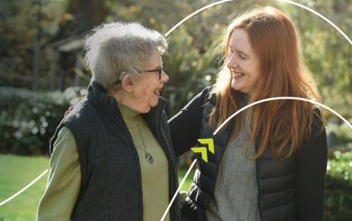Two people standing together outdoors in a garden, with one person placing an arm around the other's shoulder.