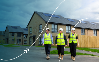 Three people wearing high-visibility clothing and hard hats walking through a residential housing development with solar panels on the roofs.