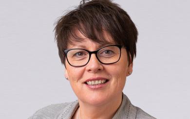 Head-and-shoulders portrait of Kathryn Fox-Rogers wearing glasses and a light grey jacket against a grey background.