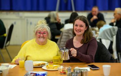 A cheerful scene in a community hall shows two women sitting at a table. The older woman on the left is wearing a bright yellow sweater and pink glasses, smiling warmly. The younger woman on the right, dressed in a dark top, is also smiling and holding a metal teapot. The table is set with plates of food, a yellow mug, and a glass of juice. In the background, other people are seated at tables, engaged in conversation.