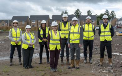 Eight people wearing white hard hats, high-visibility yellow safety vests and work boots stand on a muddy housing construction site. Newly built brick houses line the background under a cloudy sky. The group faces the camera in front of the developing residential area, with construction materials and vehicles visible around the site.