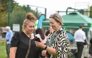 Two women are standing outdoors, engaged in conversation while looking at a smartphone. One woman, with a bun hairstyle and wearing a black top, appears to be listening intently. The other woman, with short grey hair and dressed in a patterned black and white outfit, is showing something on the phone. In the background, there are people mingling and green tents set up, indicating a social event or gathering.