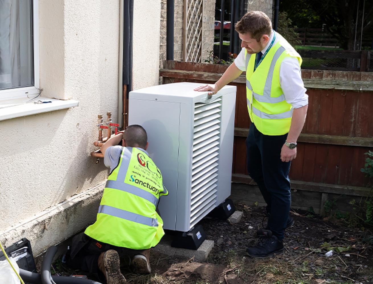 Two men wearing yellow high visibility vests work on a heating unit outside of a house