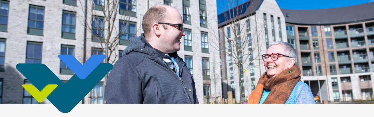 A man and a woman stand outside smiling at each other with a multi-storey building in the background 