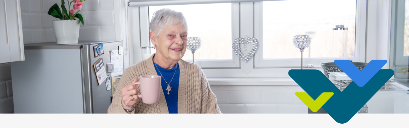 An elderly lady standing in a white kitchen holding a pink mug