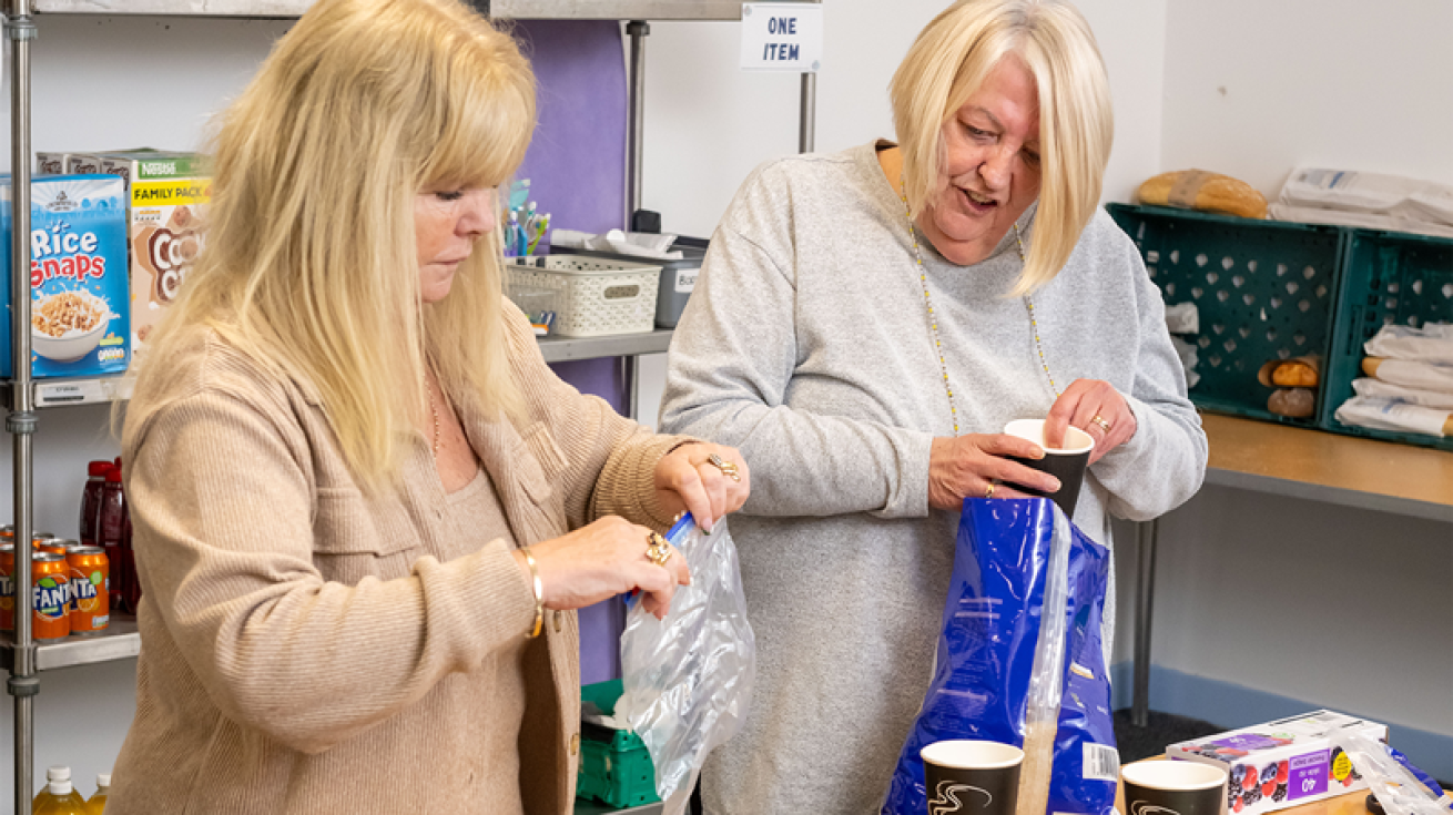 Two women standing in a room with produce stacked on shelves in the back ground. One is holding a plastic bag and the other is holding a takeaway coffee cup