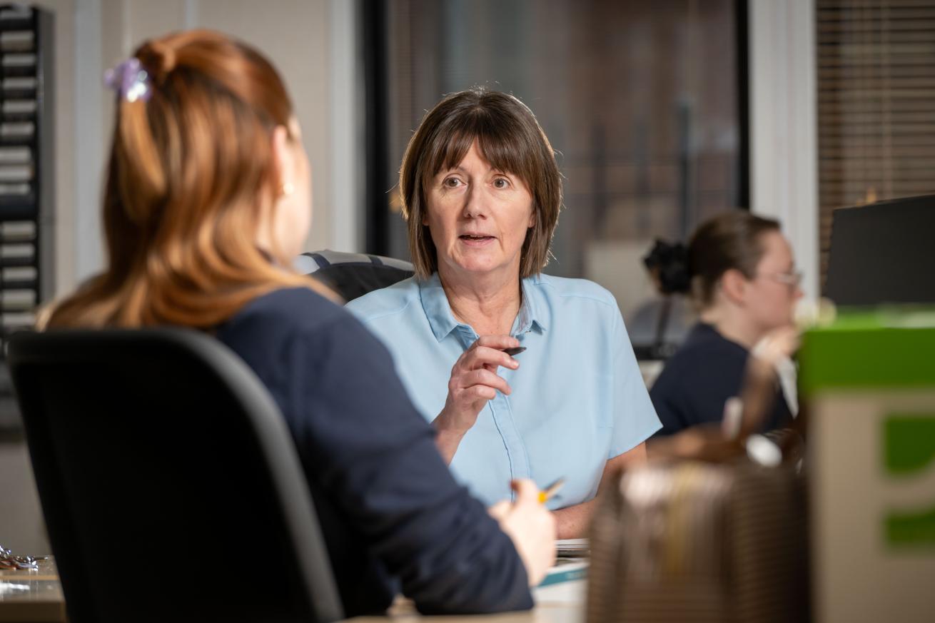 A woman sitting at an office desk speaking to another woman