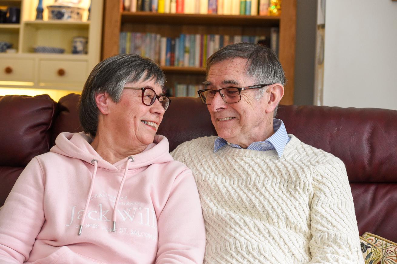 Two people wearing cosy jumpers looking into each other's eyes while sitting on a dar red sofa