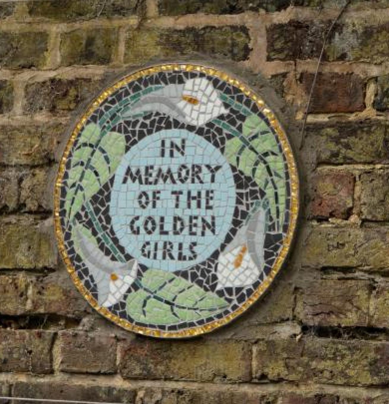 Circular mosaic on a brick wall with white flowers and green leaves, featuring the text "IN MEMORY OF THE GOLDEN GIRLS" in the center.