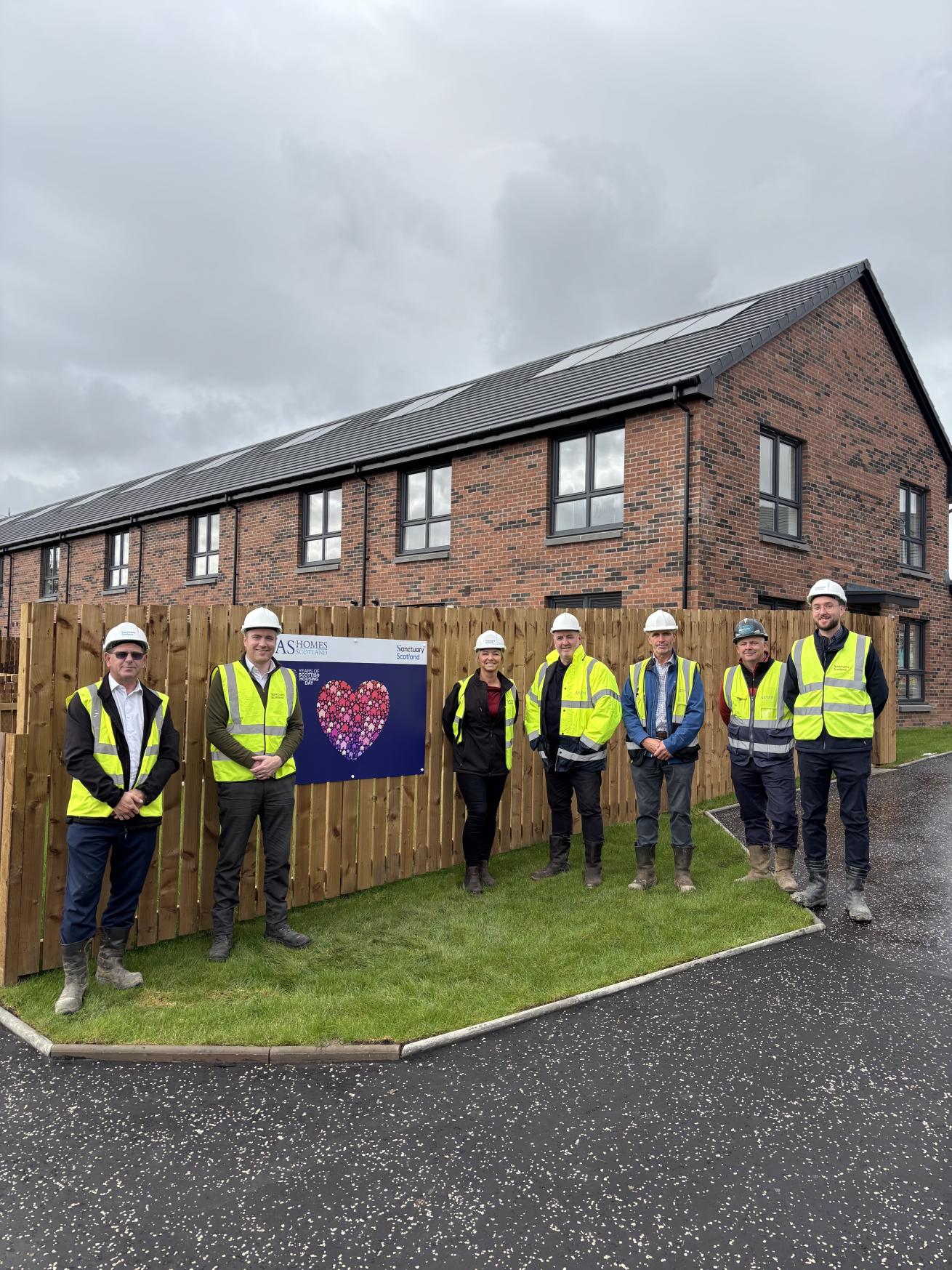 A group of people in hard hats and yellow hi-vis jackets stood in front of a row of new-build houses.