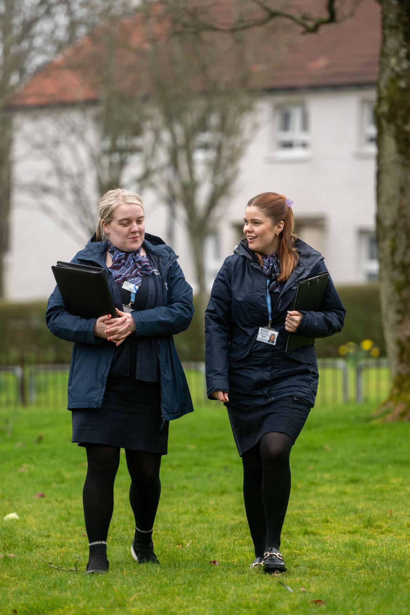 Two women walking side by side on a grassy area, both dressed in dark blue jackets and skirts.