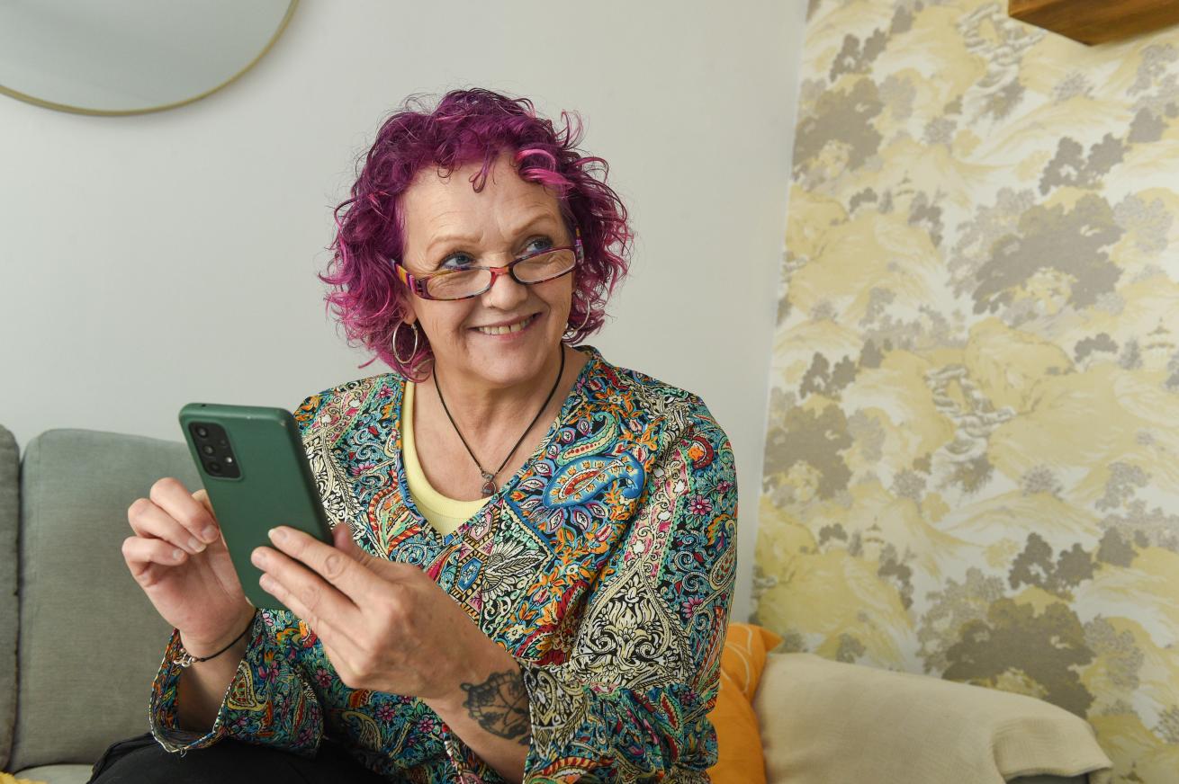 A smiling woman with curly, vibrant purple hair is sitting on a couch, holding a smartphone in her hands.
