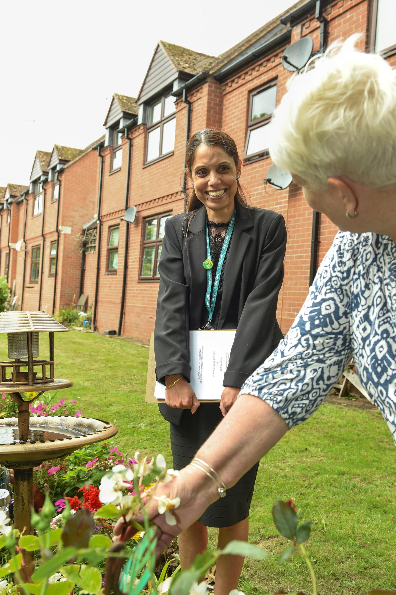 A woman in a black suit and holding a folder smiles at the camera while standing in a garden. She is interacting with another woman, who is leaning over to examine flowers.