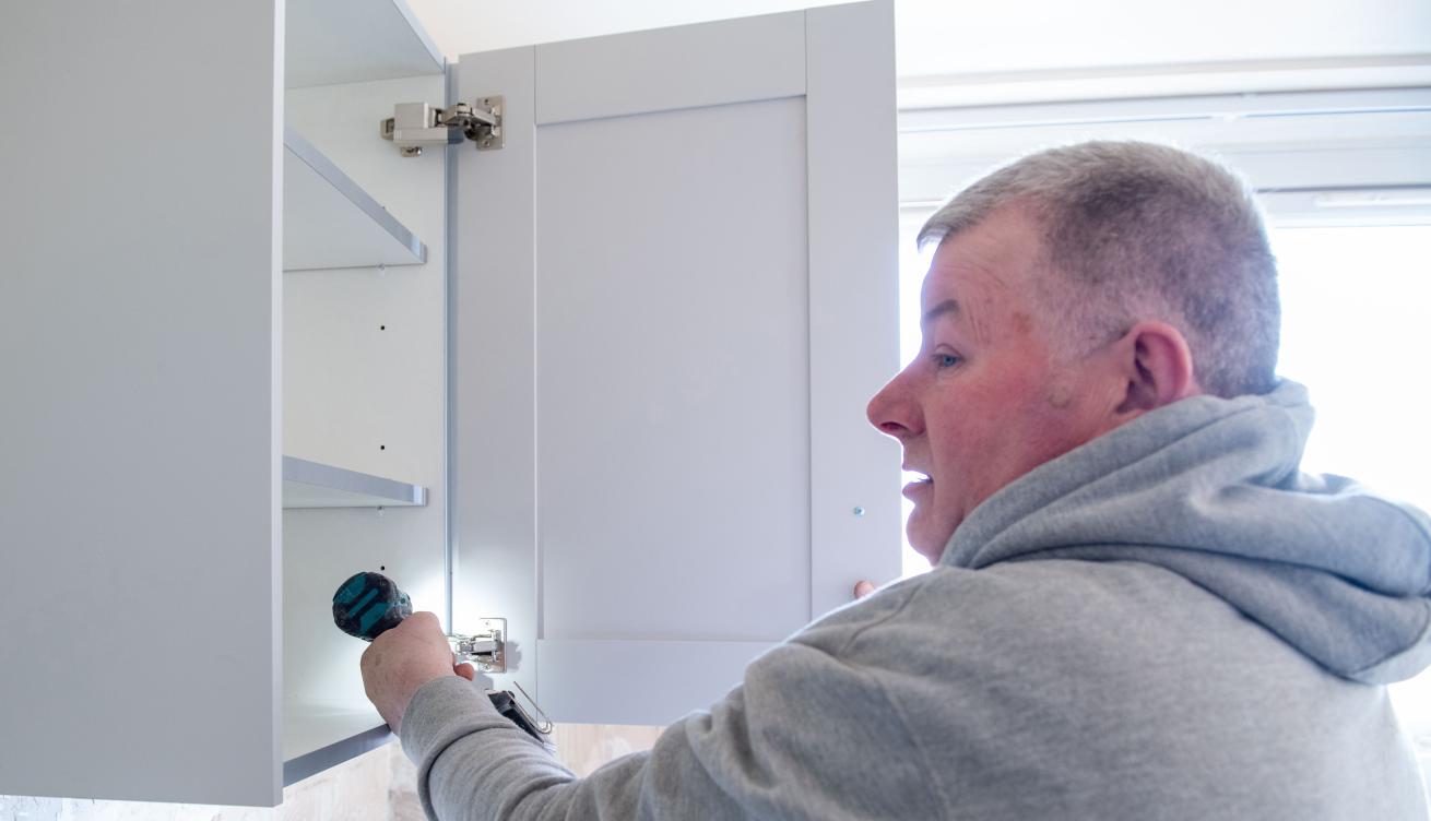 A man wearing a gray hoodie is using a power drill to install shelves inside a light gray kitchen cabinet.
