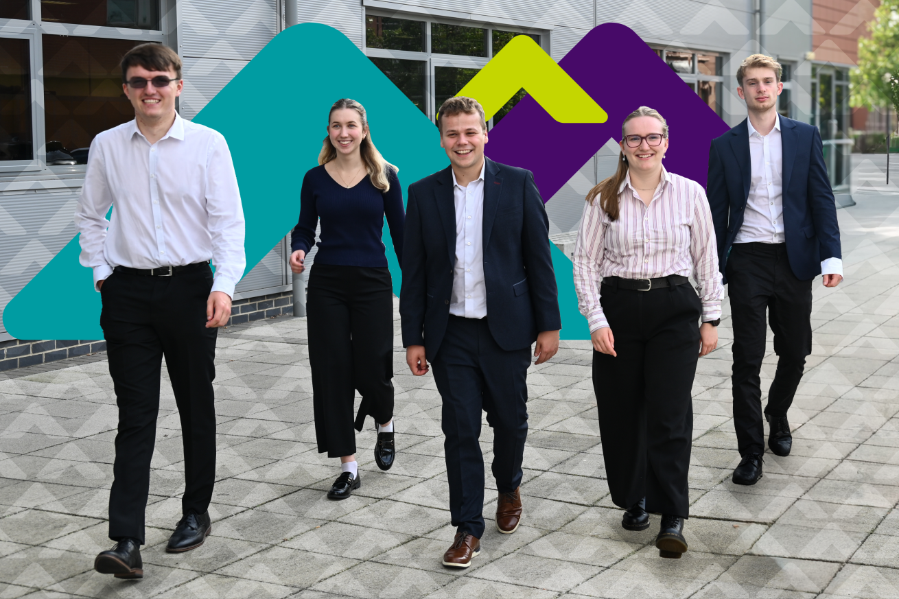 Five young professionals in business attire, with three men wearing white and dark suits, and two women in a striped blouse and a dark sweater paired with tailored pants. They are walking confidently on a paved walkway outside a modern building.