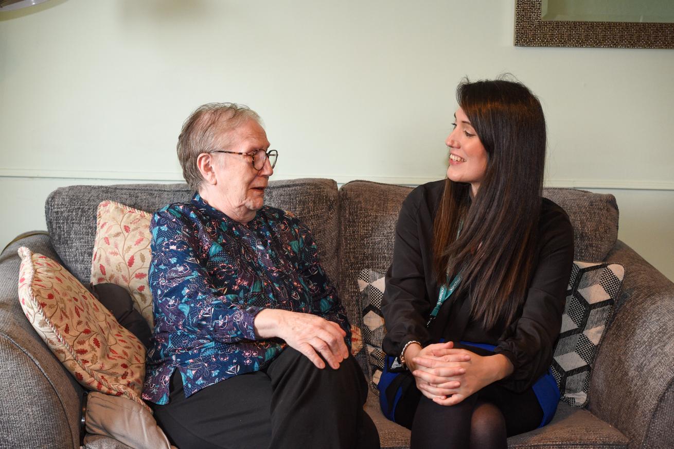 senior woman and a younger woman are sitting together on a cozy gray sofa, smiling and engaging in conversation. The older woman, wearing glasses and a patterned blouse, appears joyful, while the younger woman, dressed in a black top, is laughing. Decorative pillows are arranged on the sofa, and the background features soft lighting and a hint of wall decor, creating a warm and inviting atmosphere.