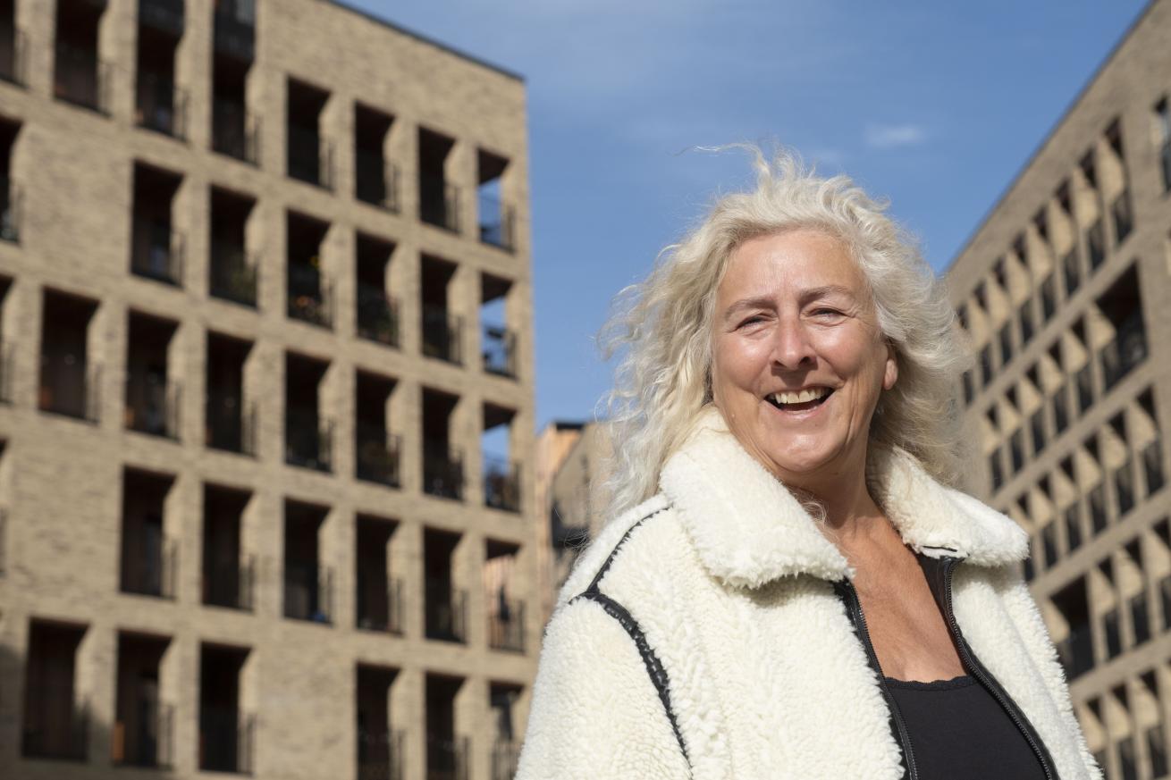 Person wearing a light-colored textured coat and dark top standing outdoors in front of modern multi-storey brick apartment buildings under a clear blue sky.