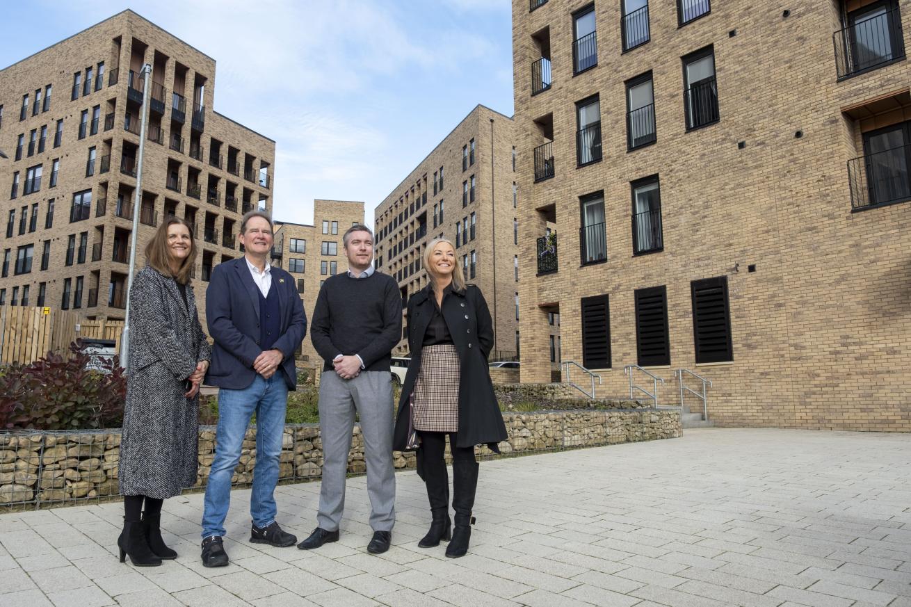 Four people standing in an outdoor courtyard area surrounded by modern multi-storey brick apartment buildings, with landscaped stone planters and paved walkways