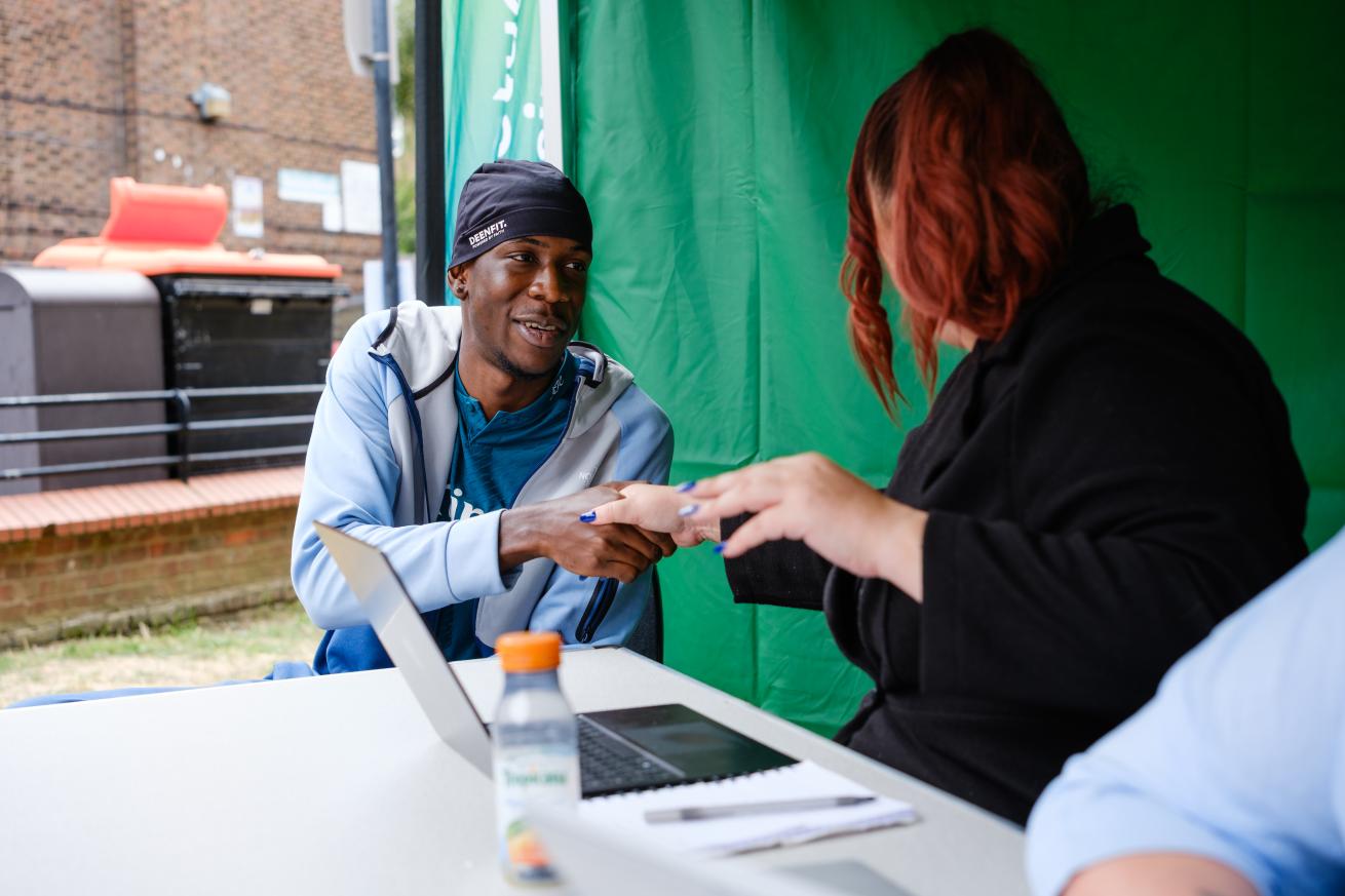 A man and a woman are seated at a table under a green tent. The man is smiling and shaking hands with the woman. A laptop and a drink are visible on the table.