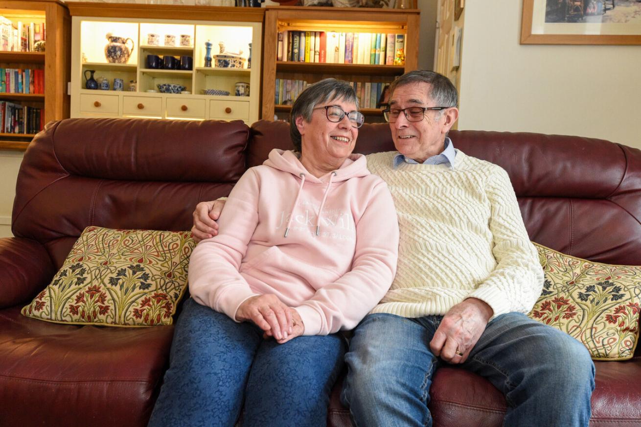 A middle-aged couple sits closely together on a comfortable sofa, smiling at each other. The woman wears a light pink hoodie and glasses, while the man is dressed in a cream-coloured sweater and glasses. They are surrounded by a cosy living room with bookshelves and decorative items in the background. The atmosphere is warm and affectionate.