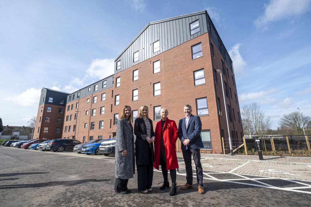 From left to right - Pat Cahill Director of Housing and Property at Sanctuary Scotland, Màiri McAllan Scottish Housing Secretary, Gill Lavety Development Director for Sanctuary Scotland, David Wylie, Managing Director, CCG Scotland. They stand infront of Adam Place a 5 story block of apartments made of brick with black panelling on the walls of the top floor.