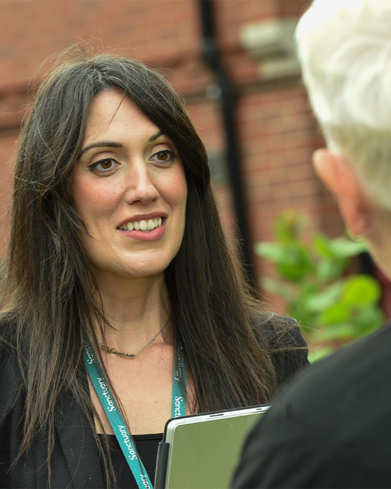 A woman with long dark hair is engaged in conversation with another person, who is partially out of frame. 