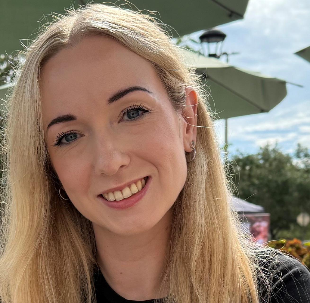 A head and shoulders photo of a woman with long blonde hair. She is wearing a smart black t-shirt and smiling towards the camera.