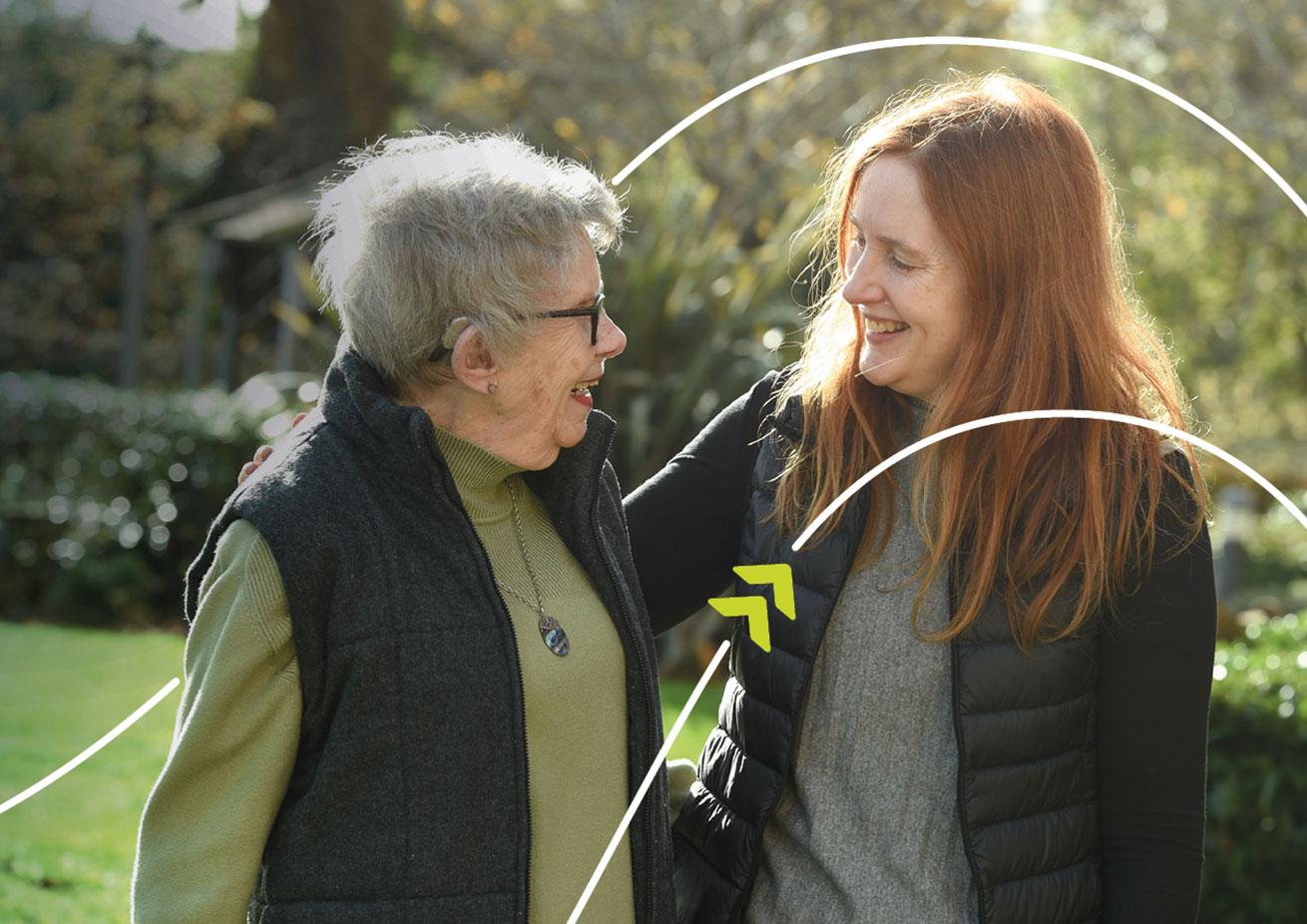 Two people standing together outdoors in a garden, with one person placing an arm around the other's shoulder.