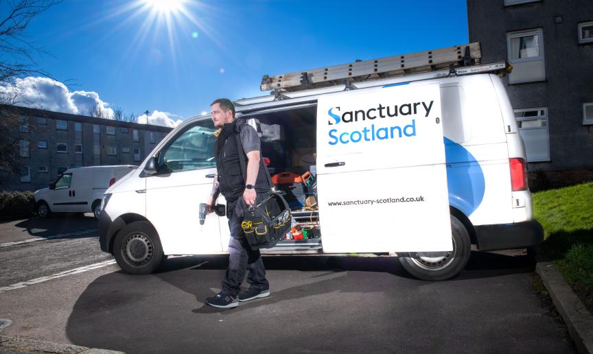 A maintenance person carrying a tool bag and a drill standing in front of a Sanctuary Scotland maintenance van