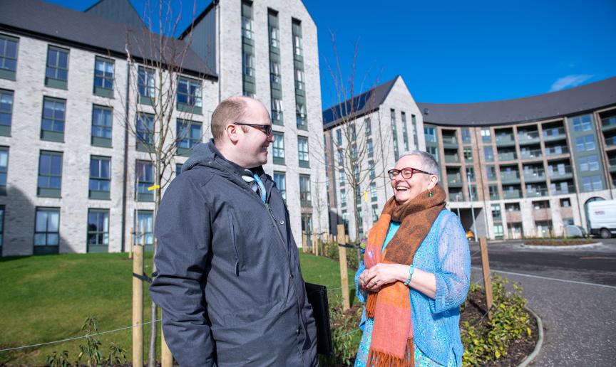 A man and a woman stand outside smiling at each other with a multi-storey building in the background 