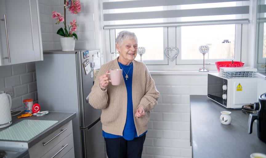 An elderly lady standing in a white kitchen holding a pink mug