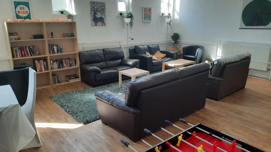 An daytime photo of the Community Living Room, showing a table and chairs, book case, sofas, coffee tables, and football table.